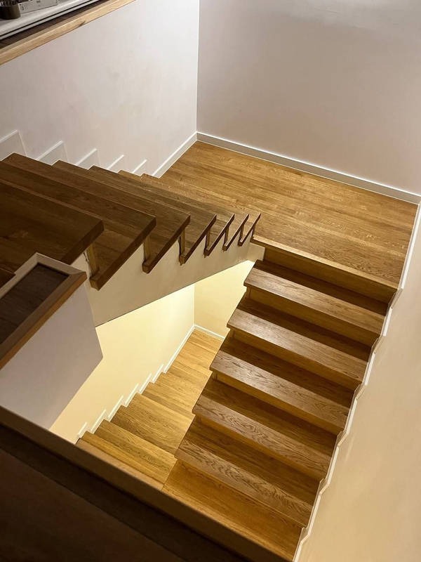 Kątowy widok z góry na drewniane schody z podstopnicami, z widocznym fragmentem drewnianej podłogi i białymi ścianami. Naturalne oświetlenie.