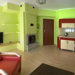 Wnętrze salonu z aneksem kuchennym po malowaniu ścian na odcień limonki. Widoczny kominek z szarego kamienia, czerwona sofa i zabudowa kuchenna w kolorze czerwieni.