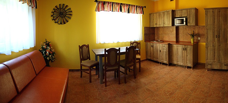 Żółte wnętrze pokoju z aneksem kuchennym: drewniane meble, stół z krzesłami, pomarańczowa sofa, dekoracje ścienne. Widok na wykończone wnętrze.