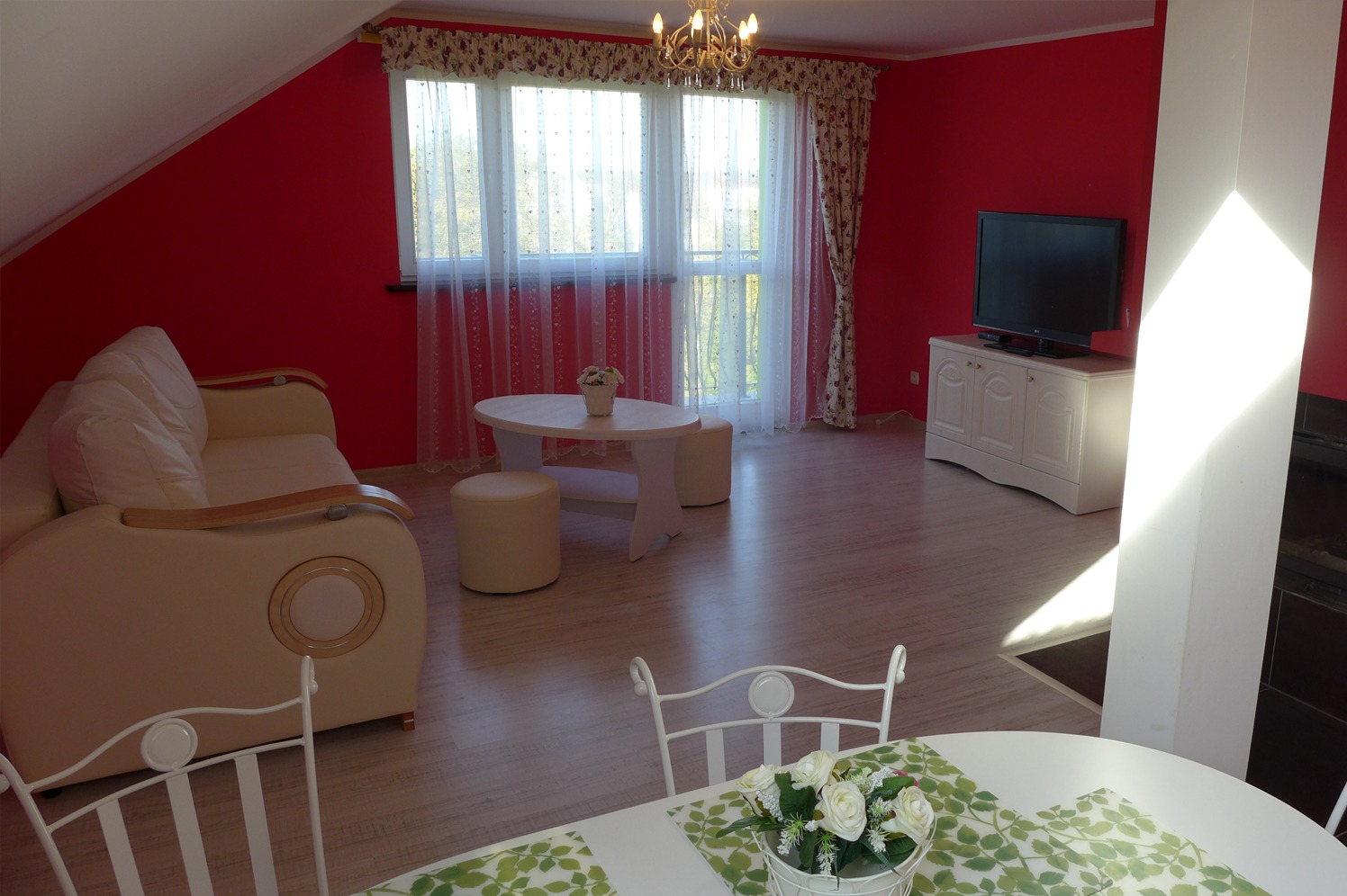 Czerwony salon na poddaszu: beżowa sofa, białe meble, okno z firankami. Widok zza stołu z bukietem kwiatów, podkreślający przytulny charakter wnętrza.