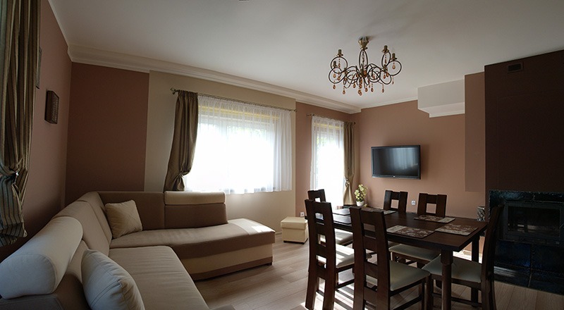 Jasny salon z brązowymi ścianami, narożną sofą i stołem jadalnym. Dekoracyjny żyrandol, zasłony w oknach i kominek dodają przytulnego charakteru wnętrzu.