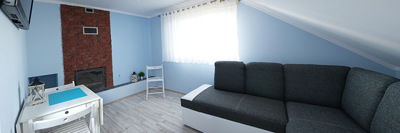 Błękitny pokój na poddaszu z ceglanym kominkiem, białymi meblami i szarą sofą. Okno z białymi firankami. Całość w jasnych, pastelowych barwach.