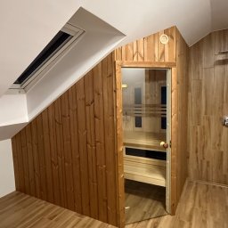 Timberhaus - Sauna wkomponowana w skos dachu, wykończona drewnem, z przeszklonymi drzwiami. Widoczne drewniane ławy wewnątrz. Podłoga wyłożona panelami imitującymi drewno.