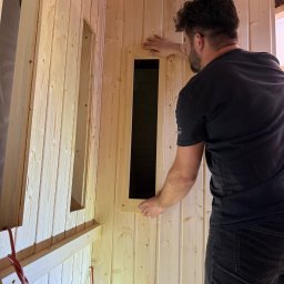 Timberhaus - Montaż drewnianej boazerii w saunie z oknami. Widoczny fragment instalacji elektrycznej i wełna mineralna. Fachowiec w trakcie pracy.