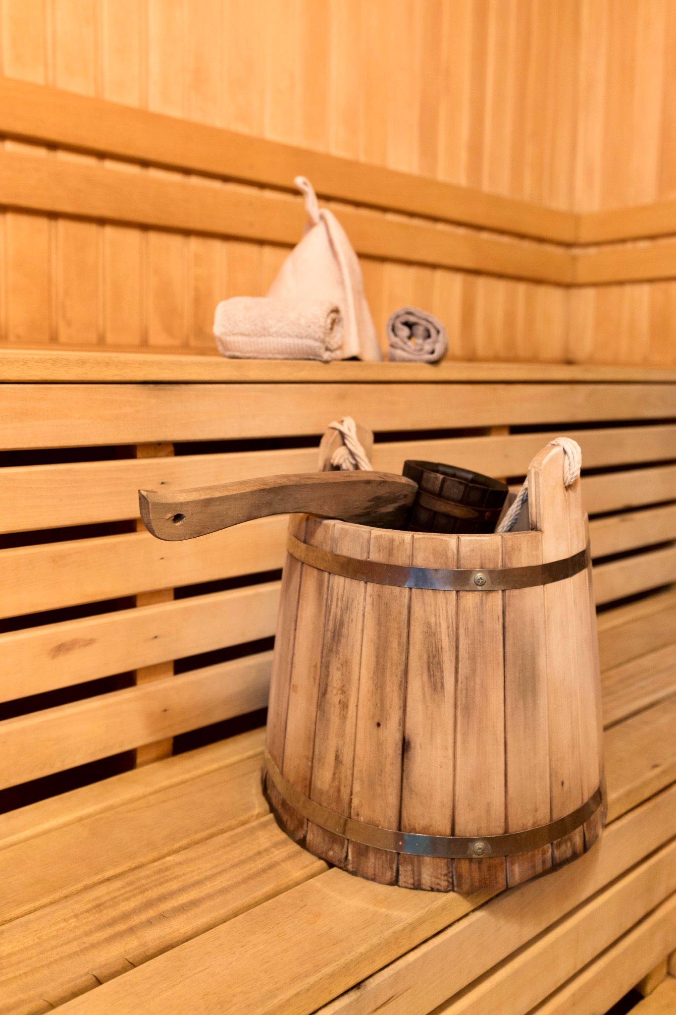 Drewniane wiadro z chochlą w saunie, obok ręcznik i czapka. Elementy wyposażenia sauny na tle drewnianych desek. Jasne, ciepłe oświetlenie.