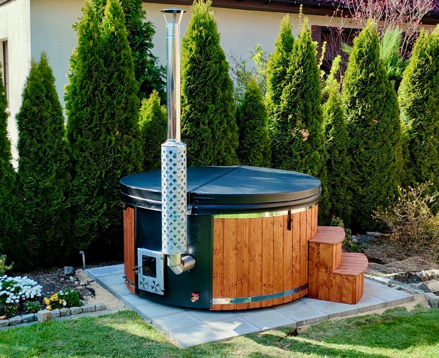 Owalna, drewniana balia ogrodowa z czarnym wykończeniem i metalowym kominem, otoczona tujami i zielonym trawnikiem, na betonowej wylewce. Schodki z boku.