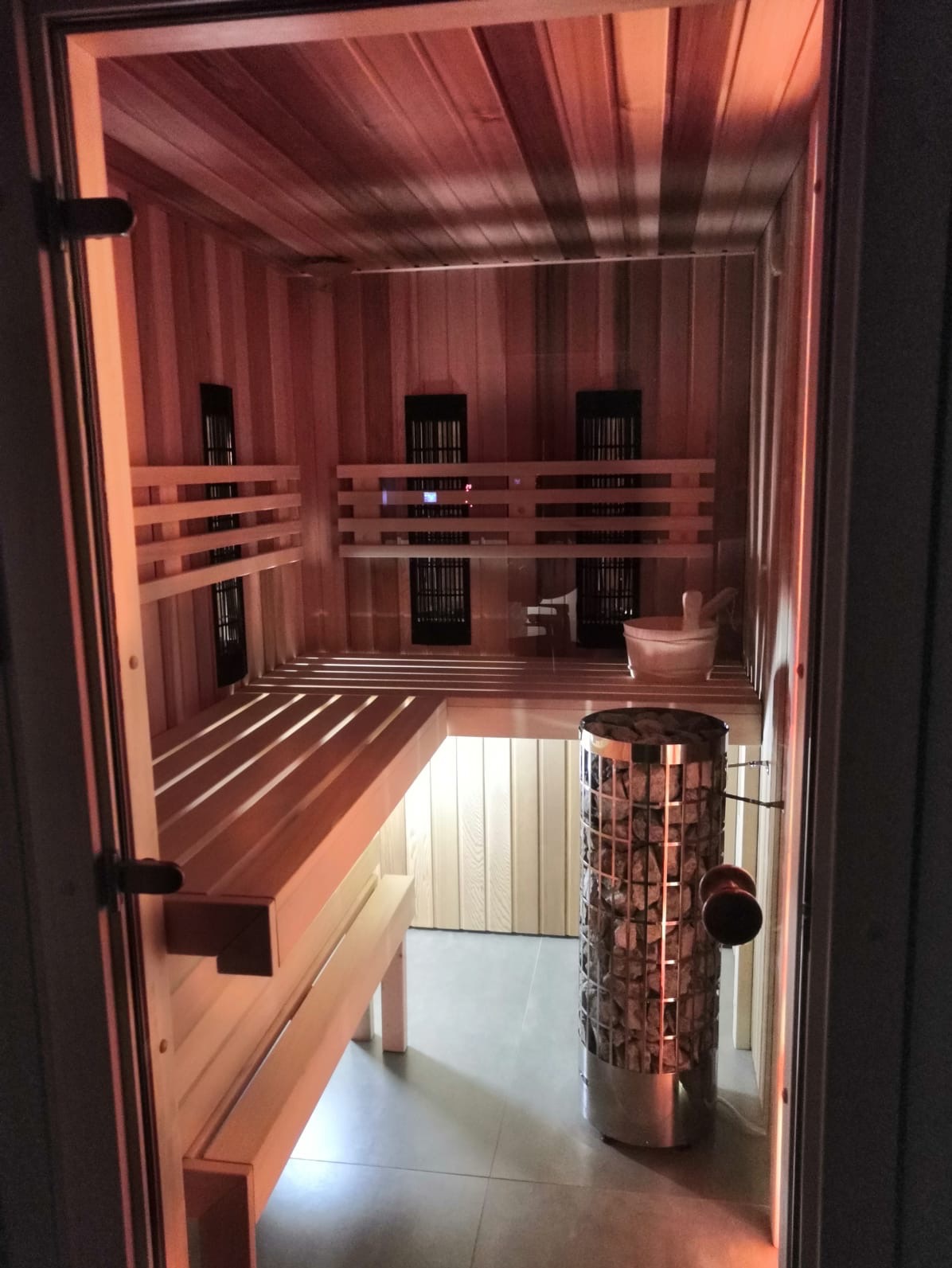 Wnętrze sauny z drewnianymi ławkami, piecem z kamieniami i oświetleniem w ciepłych tonach, widoczne przez otwarte drzwi. Detal: wiadro i chochla na półce.