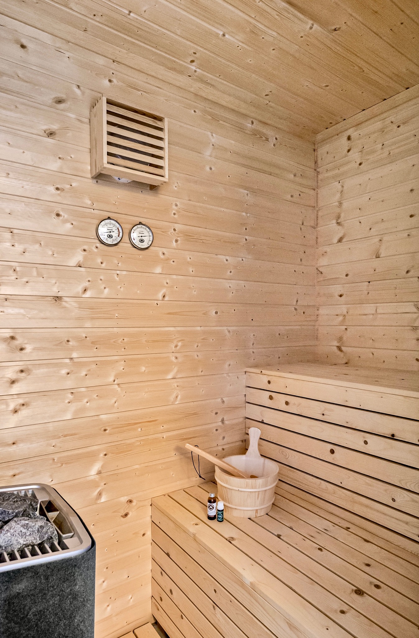 Jasne drewno w saunie: ławki, ściany, sufit. Na ławce wiadro z chochlą i olejki eteryczne. Piec z kamieniami w rogu. Dwa termometry na ścianie.