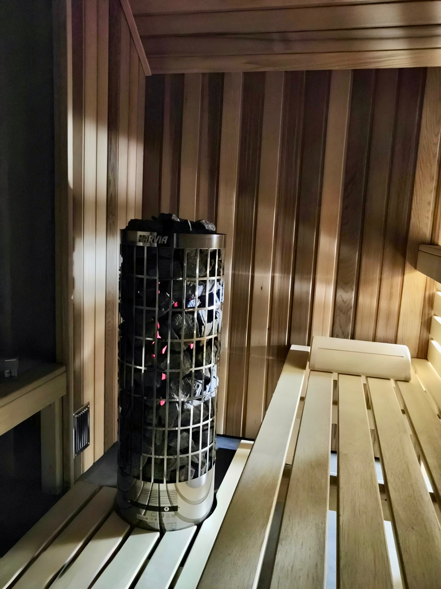 Wnętrze sauny z drewnianym wykończeniem, piecem Harvia wypełnionym kamieniami i ławkami. Ciepłe oświetlenie tworzy relaksującą atmosferę.