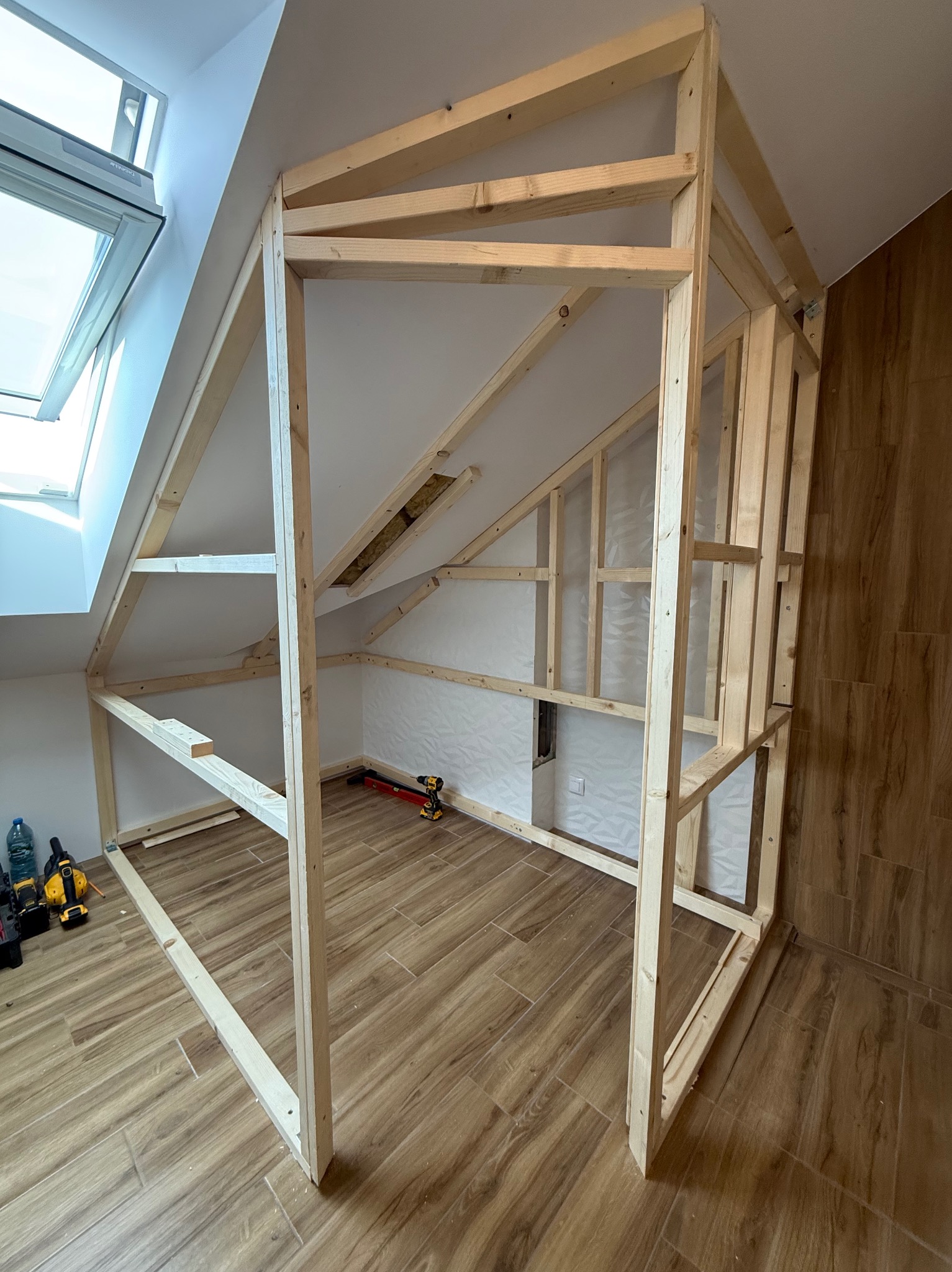 Drewniana konstrukcja szkieletowa w pokoju na poddaszu z oknem dachowym. Widoczne narzędzia i butelka wody. Podłoga wyłożona płytkami imitującymi drewno.