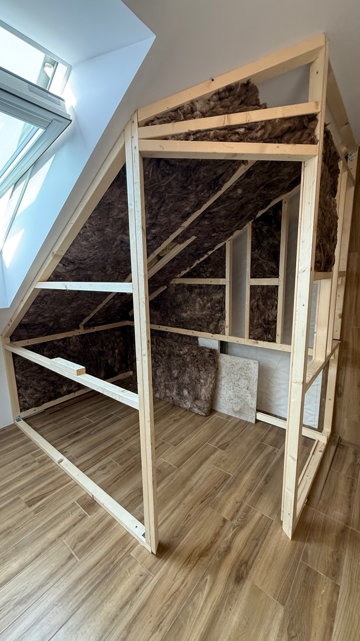 Drewniana konstrukcja szkieletowa z izolacją z wełny mineralnej na poddaszu, widok z dołu, okno dachowe w tle, płytki podłogowe imitujące drewno.