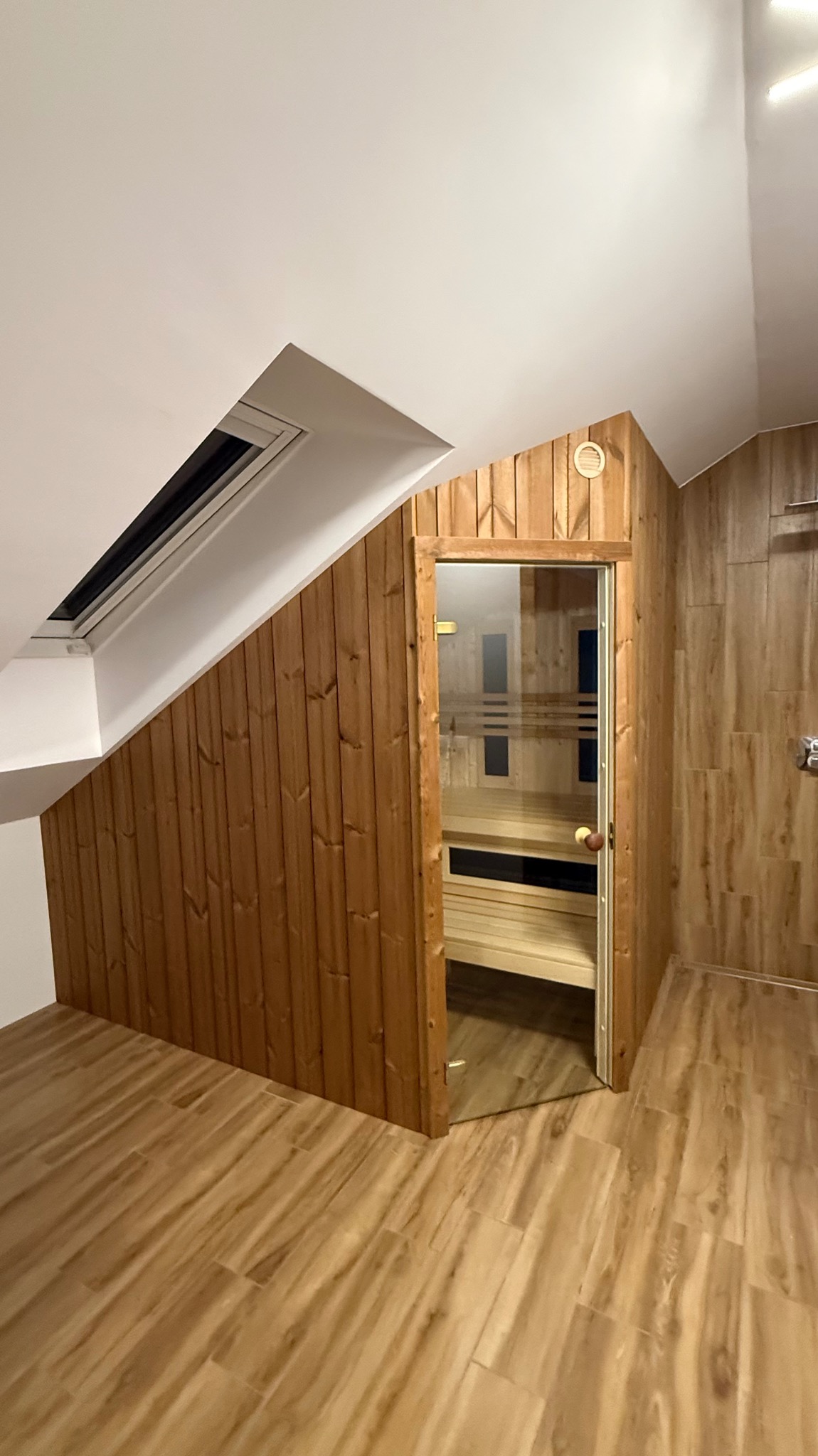 Sauna wkomponowana w skos dachu, wykończona drewnem, z przeszklonymi drzwiami. Widoczne drewniane ławy wewnątrz. Podłoga wyłożona panelami imitującymi drewno.