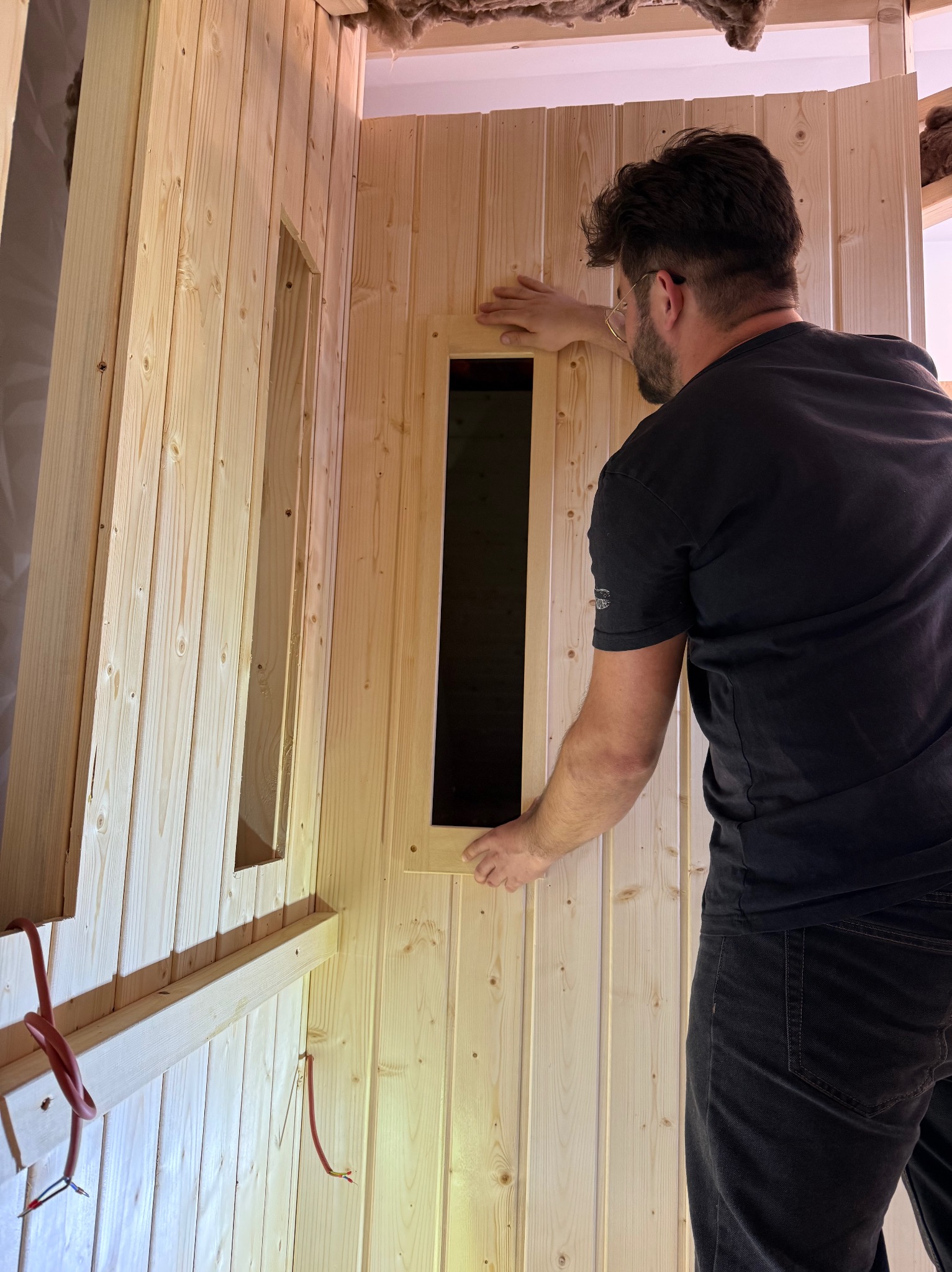 Montaż drewnianej boazerii w saunie z oknami. Widoczny fragment instalacji elektrycznej i wełna mineralna. Fachowiec w trakcie pracy.