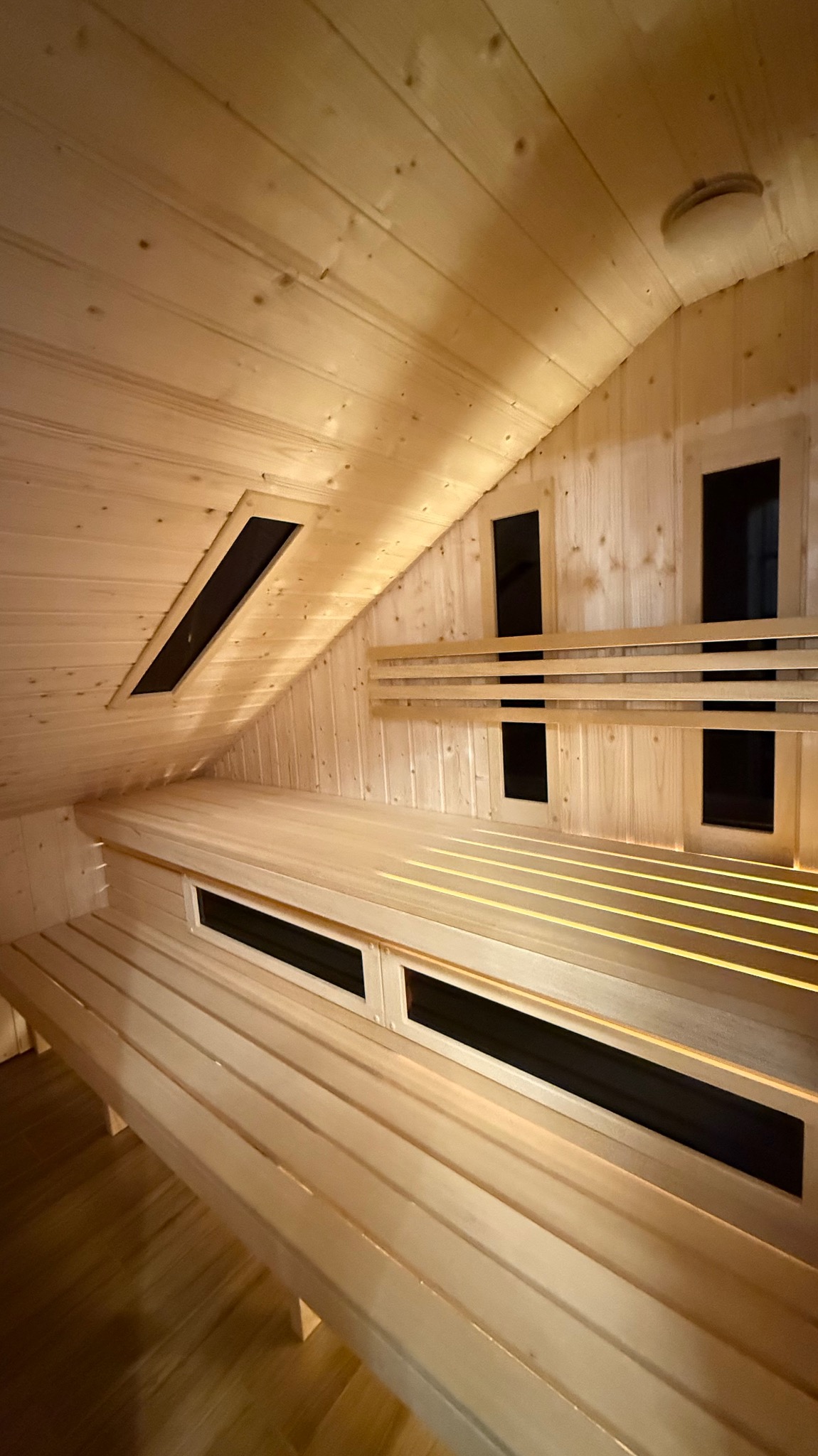 Jasne drewno w saunie: ławy, ściany i sufit z oknami wentylacyjnymi. Ukośny sufit i ciepłe oświetlenie tworzą relaksującą atmosferę.