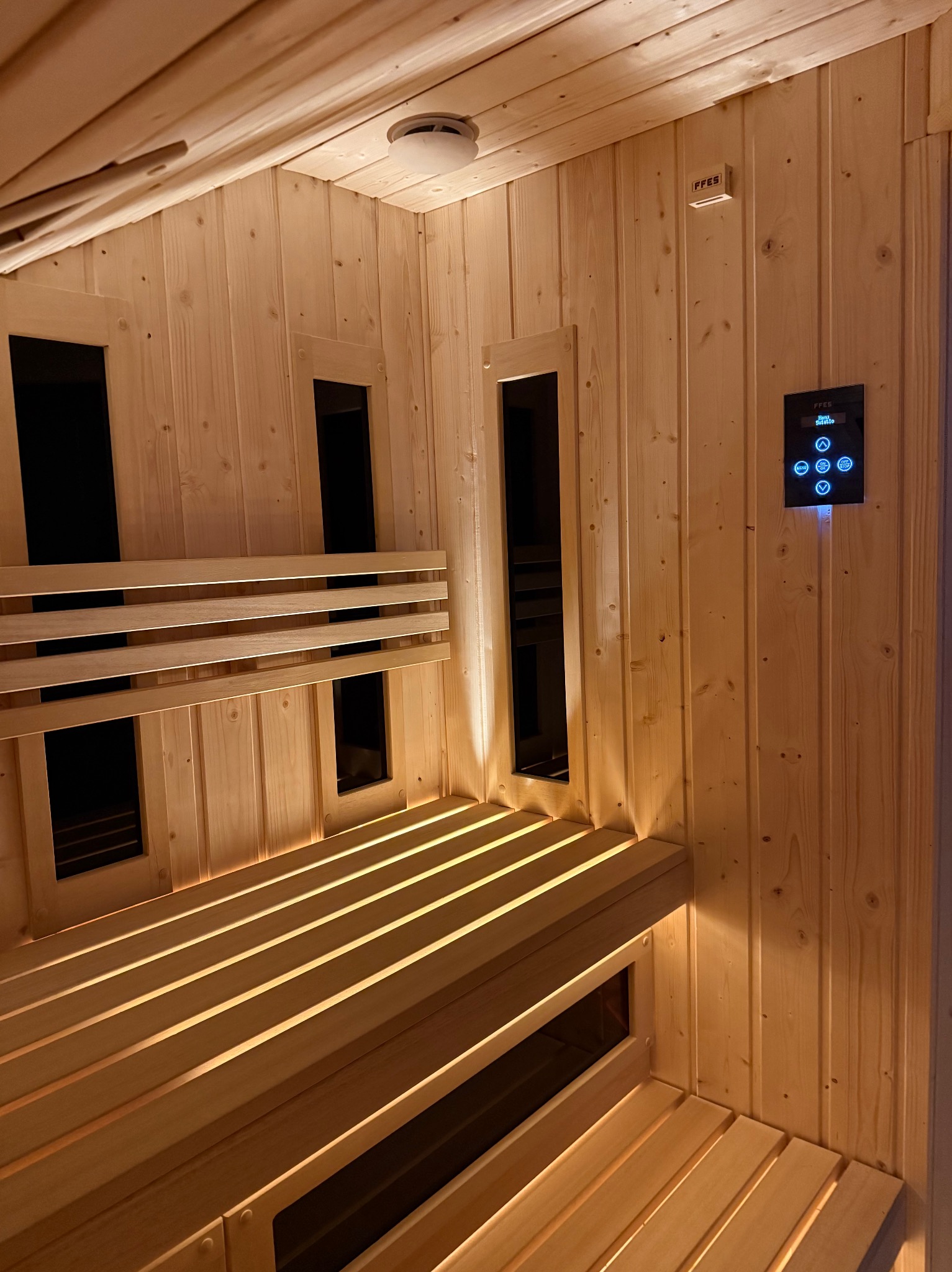 Jasne drewno w saunie z podświetlanymi ławkami i panelem sterowania. Widoczne małe okienka w ścianach. Ciepłe oświetlenie tworzy relaksującą atmosferę.