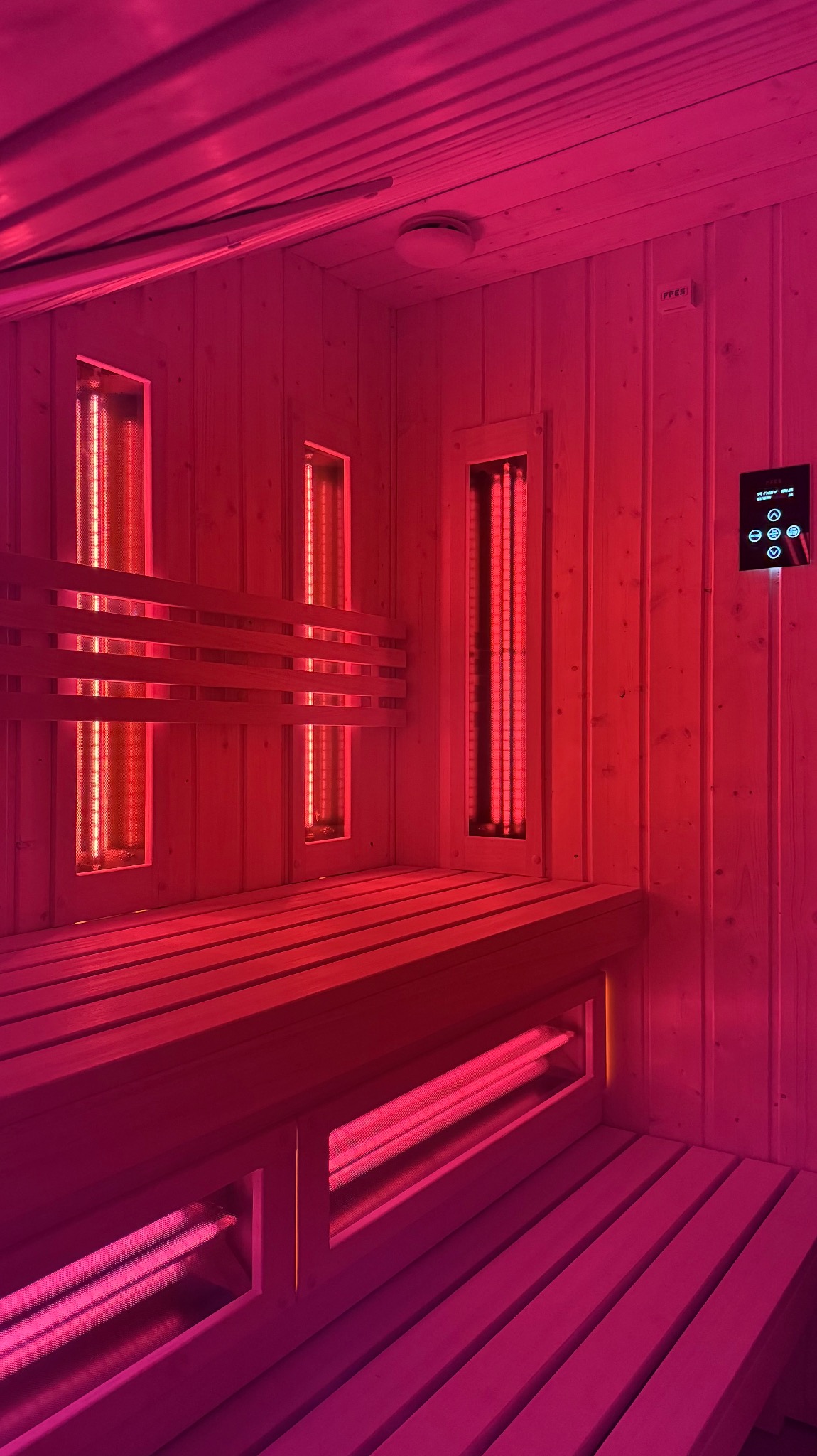 Wnętrze sauny infrared z drewnianą ławą i panelami grzewczymi, oświetlone intensywnym czerwonym światłem. Widoczny panel sterowania na ścianie.