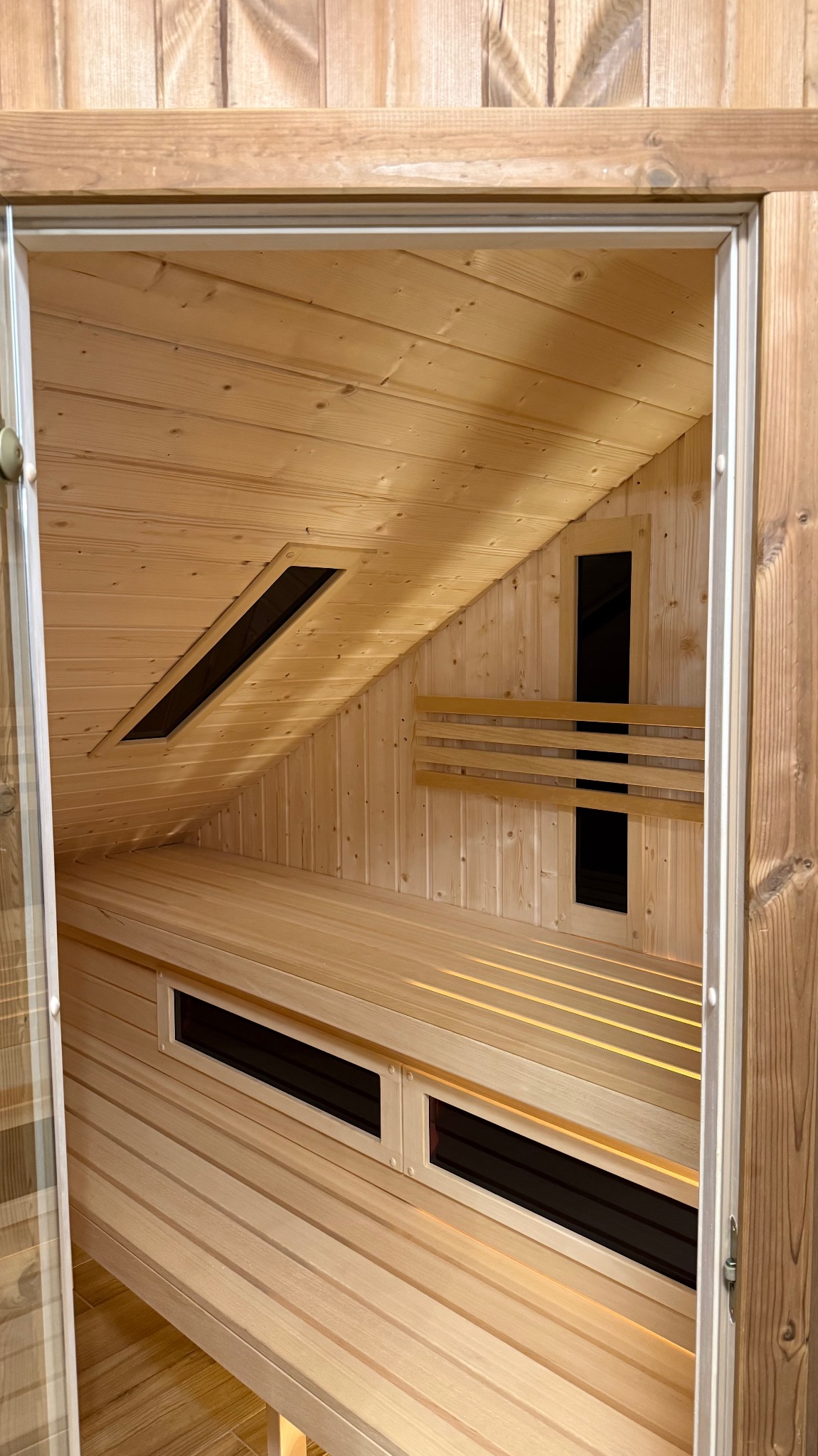 Jasne drewno w saunie: ławki, ściany i sufit z ukośnym oknem. Widok zza otwartych drzwi. Minimalistyczny design, ciepłe oświetlenie.