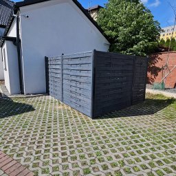 Dorbudex Paweł Dorabiała - Szara, drewniana osłona śmietnikowa na betonowej ażurowej nawierzchni z rosnącą trawą, obok białego budynku z czarnym dachem, w słoneczny dzień.