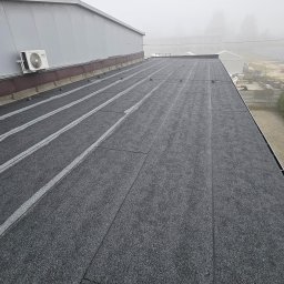 Dorbudex Paweł Dorabiała - Szczegółowe ujęcie płaskiego dachu pokrytego czarną papą, z widocznym systemem odprowadzania wody i klimatyzatorem na tle zamglonego krajobrazu.