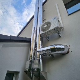 Dorbudex Paweł Dorabiała - Błyszczące, stalowe kominy na elewacji domu, obok klimatyzator. Widoczne fragmenty dachu, okno i błękitne niebo z chmurami. Ujęcie z dołu, podkreślające wysokość kominów.