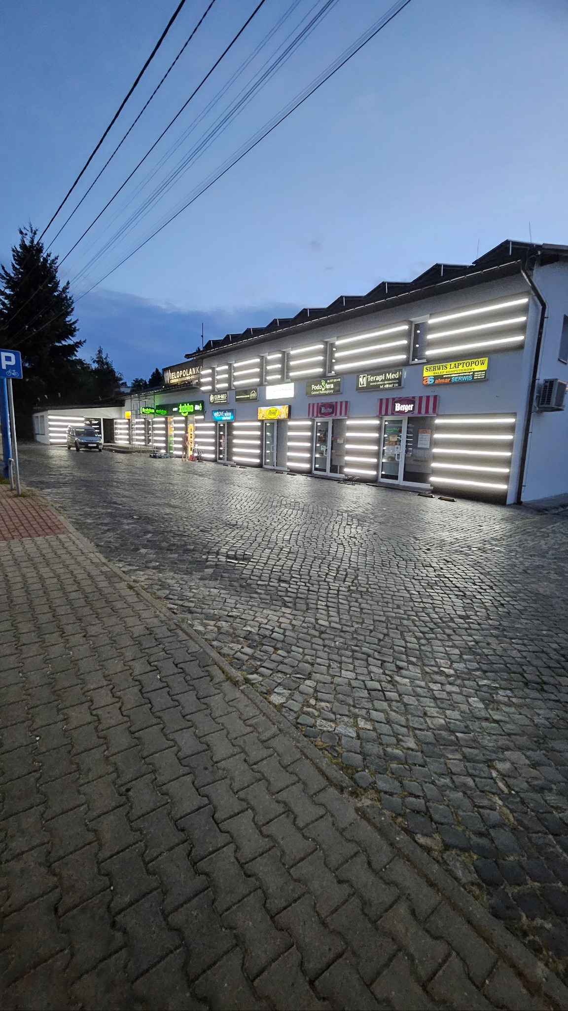 Fasada budynku usługowego z podświetlonymi lokalami i brukowanym chodnikiem o zmierzchu. Widoczne szyldy różnych firm, oświetlenie LED i zaparkowany samochód.
