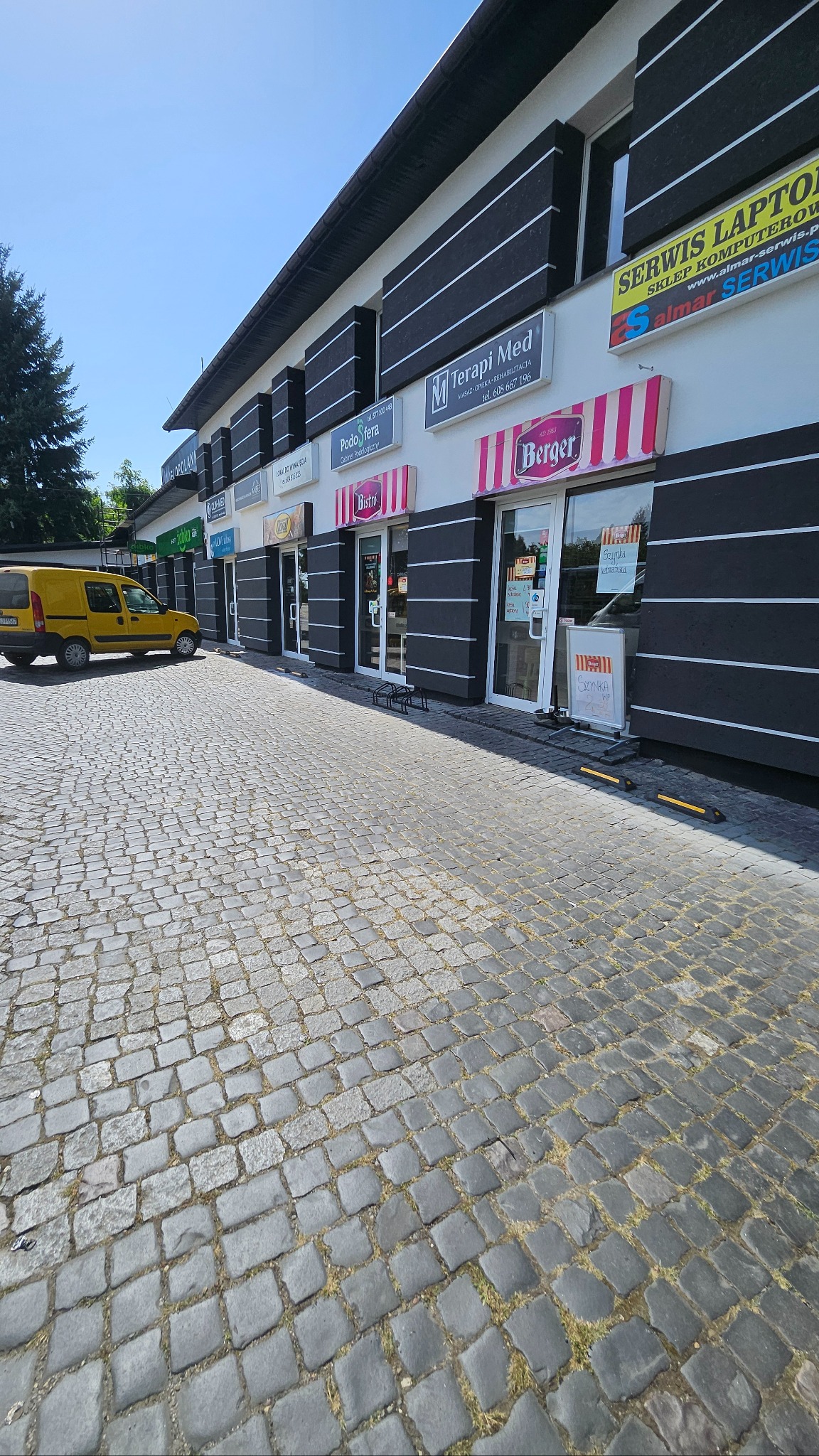 Fasada budynku usługowego z lokalami handlowymi: Bistro, Terapi Med, Berger i inne. Żółty samochód dostawczy zaparkowany obok. Kostka brukowa na pierwszym planie.