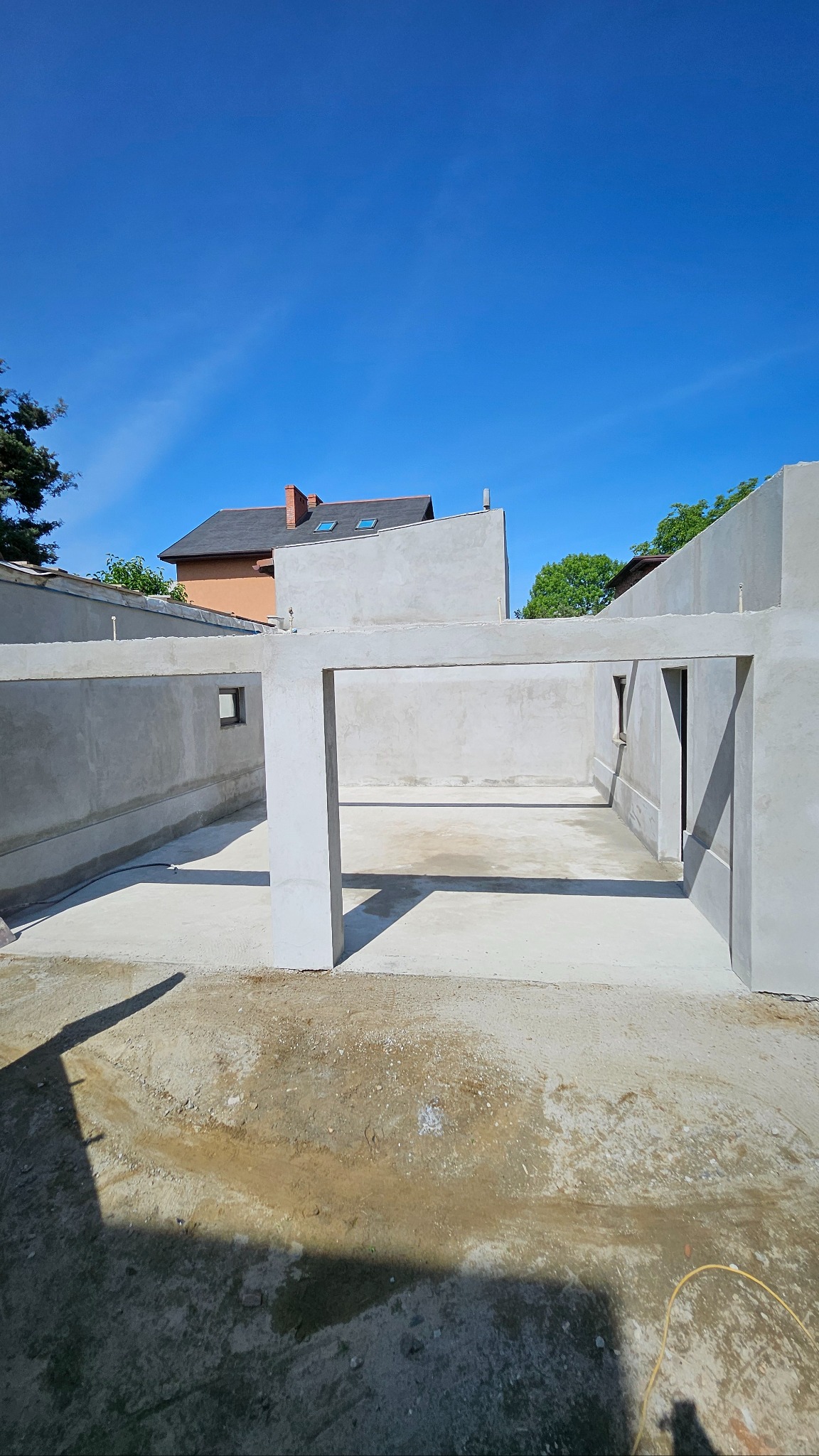 Surowa konstrukcja budynku z betonu w słoneczny dzień, widok z zewnątrz. W tle fragment dachu sąsiedniego budynku i błękitne niebo. Widoczne ślady budowy na ziemi.