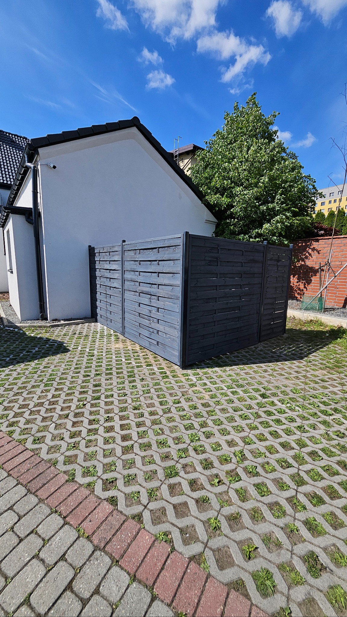 Szara, drewniana osłona śmietnikowa na betonowej ażurowej nawierzchni z rosnącą trawą, obok białego budynku z czarnym dachem, w słoneczny dzień.