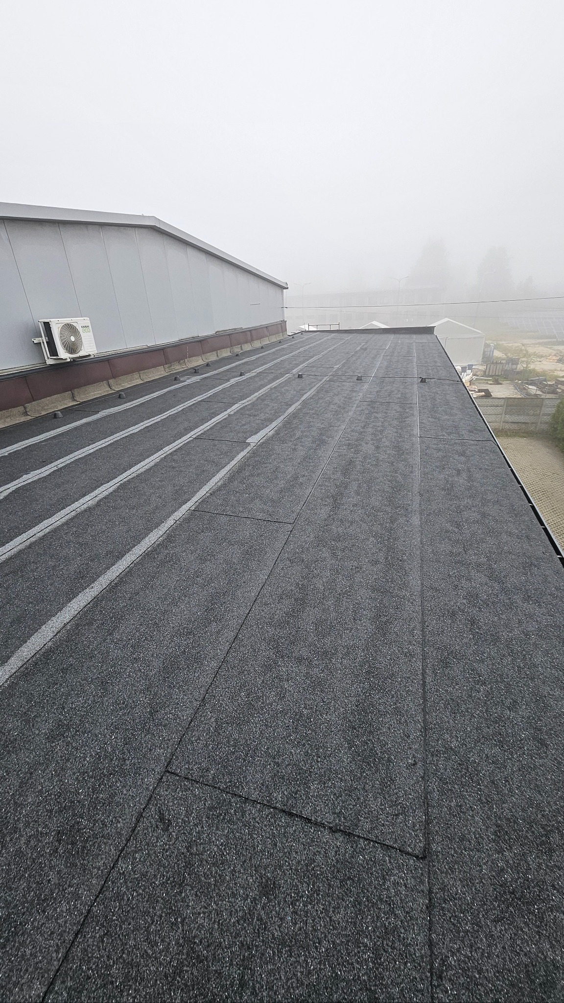 Szczegółowe ujęcie płaskiego dachu pokrytego czarną papą, z widocznym systemem odprowadzania wody i klimatyzatorem na tle zamglonego krajobrazu.