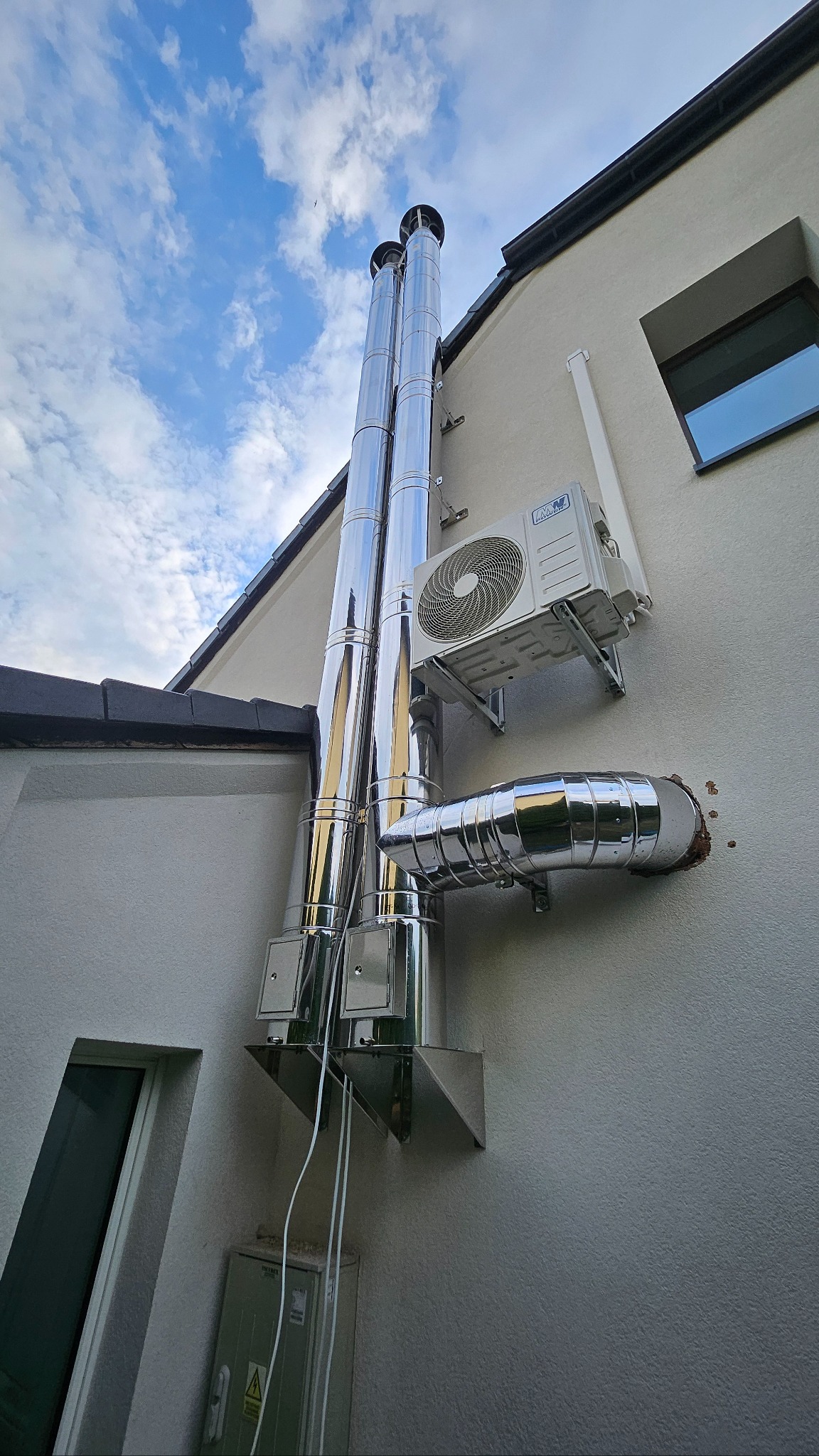 Błyszczące, stalowe kominy na elewacji domu, obok klimatyzator. Widoczne fragmenty dachu, okno i błękitne niebo z chmurami. Ujęcie z dołu, podkreślające wysokość kominów.