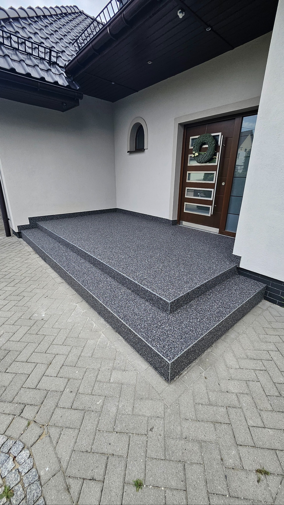 Wykończone żywicą schody wejściowe do domu z ciemnymi drzwiami i wieńcem. Nowoczesna aranżacja z szarą posadzką żywiczną na zewnątrz budynku.