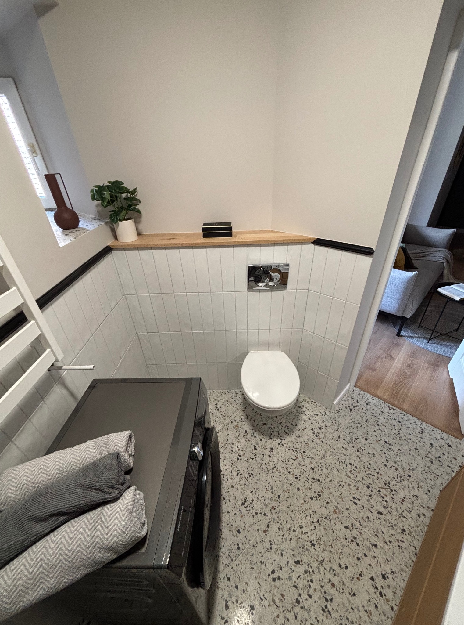 Nowoczesna toaleta w łazience po remoncie: białe, pionowe płytki, blat z drewna, pralka z ręcznikami, podłoga lastryko. Widok na salon przez otwarte drzwi.