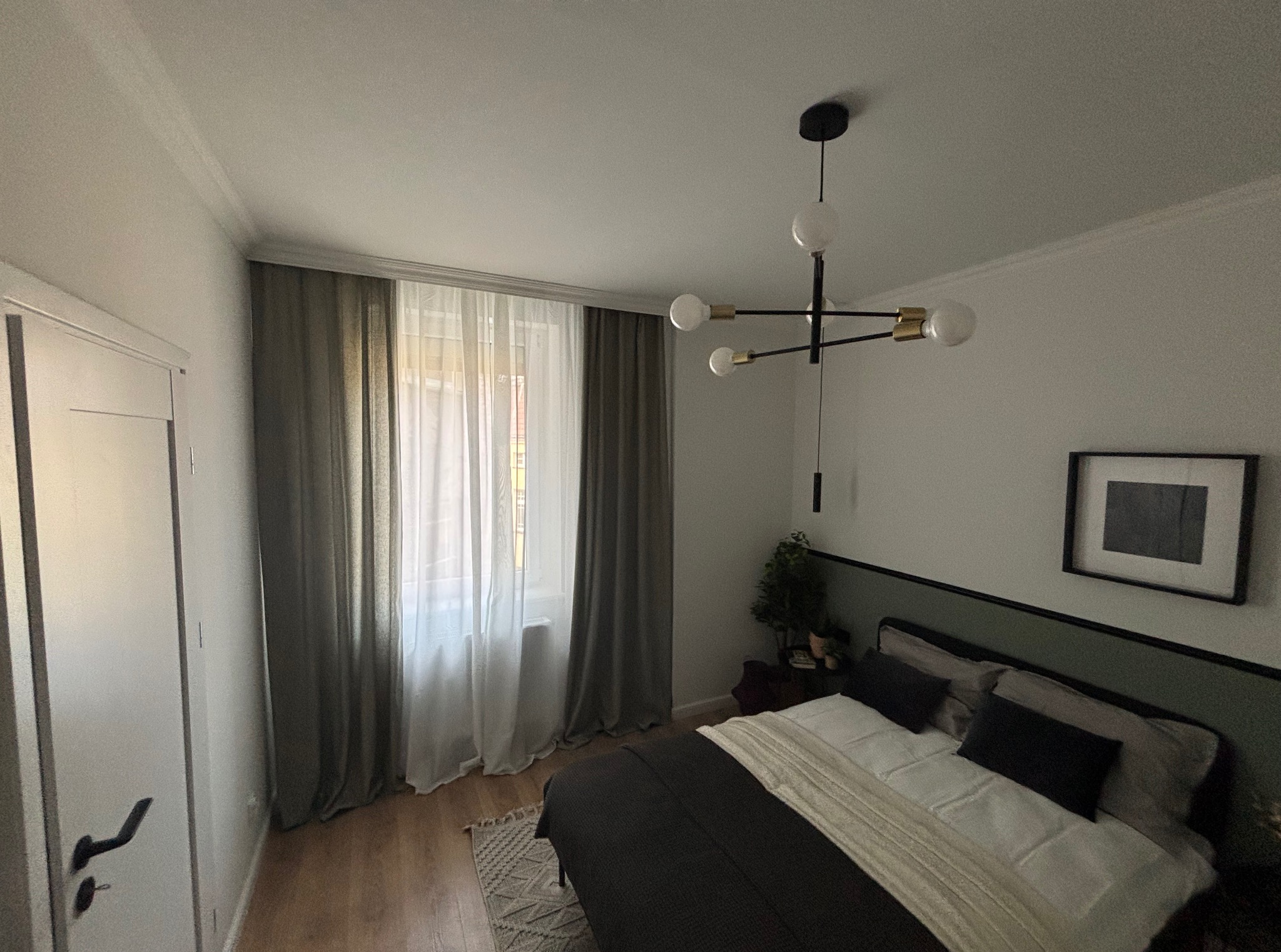 Nowoczesna sypialnia z łóżkiem, zasłonami i lampą sufitową. Ściany w odcieniach bieli, ciemne zasłony i rama łóżka. Minimalistyczny wystrój wnętrza.