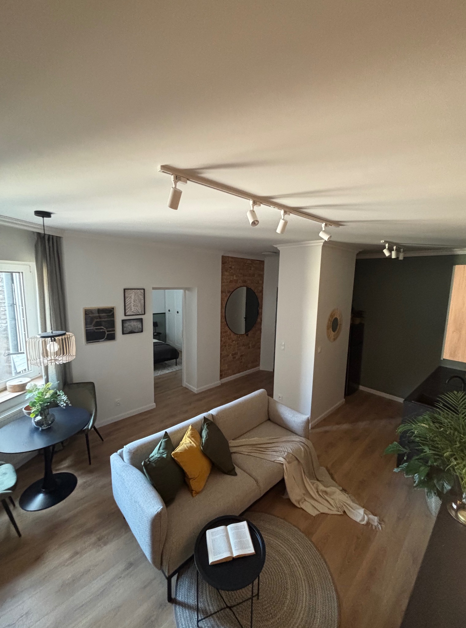 Ujęcie z góry na jasny salon z kanapą, stolikiem kawowym z książką, okrągłym stołem i roślinami. Widoczne oświetlenie sufitowe i dekoracje ścienne.