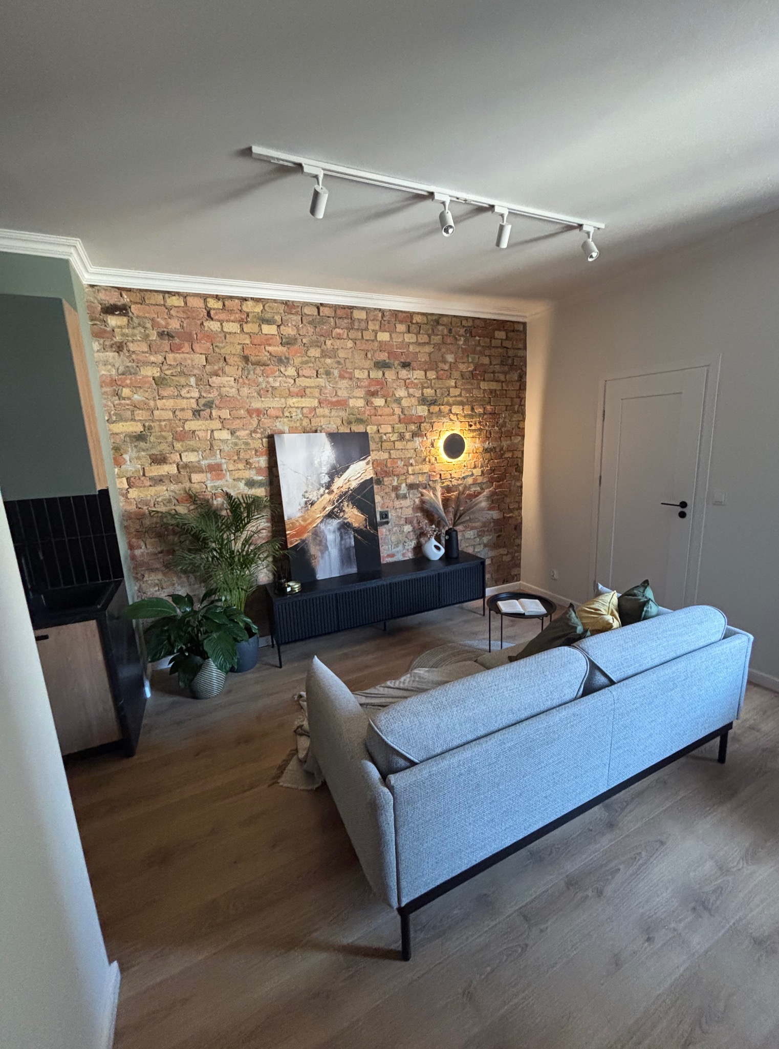 Salon z ceglaną ścianą, szarą sofą i drewnianą podłogą. Na ścianie obraz i okrągła lampa. Widoczne rośliny doniczkowe i czarna szafka RTV.