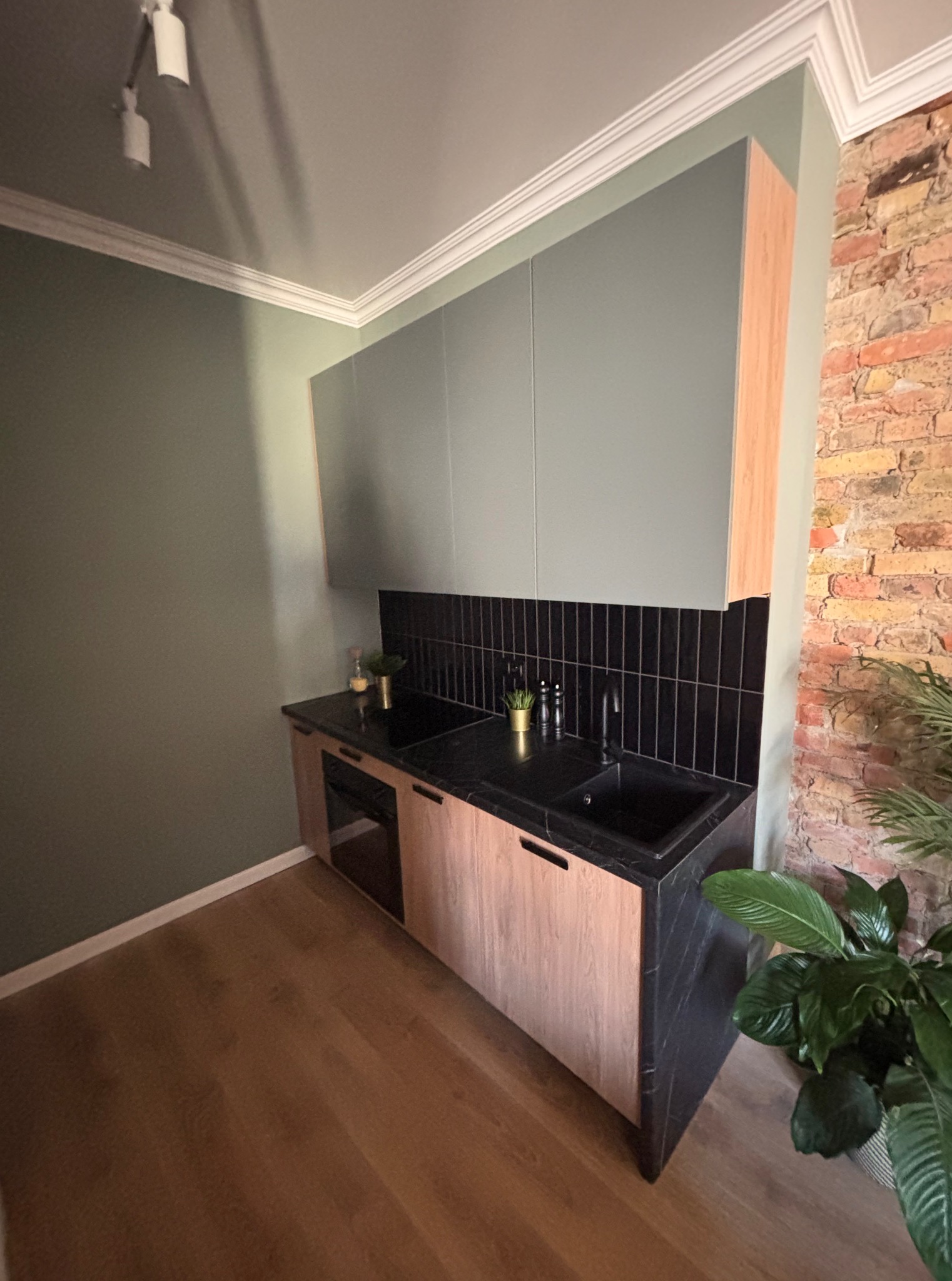 Nowoczesna, kompaktowa kuchnia z drewnianymi szafkami, czarnym blatem i pionową czarną glazurą. Ściana z cegły i zielona ściana. Podłoga drewniana.