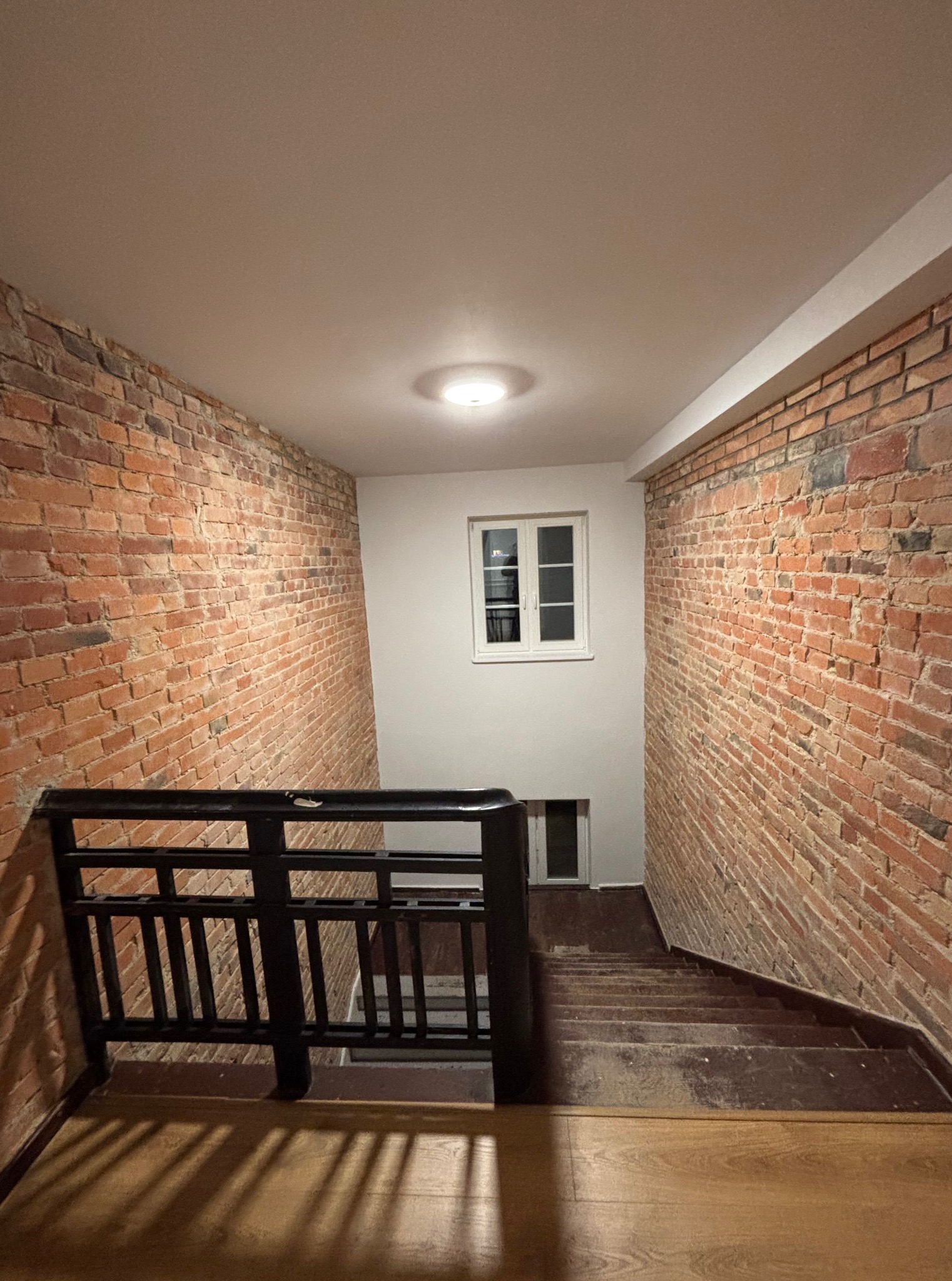 Klatka schodowa z ceglanymi ścianami i drewnianymi schodami. Widoczne okno na podeście i czarna balustrada. Oświetlenie sufitowe.