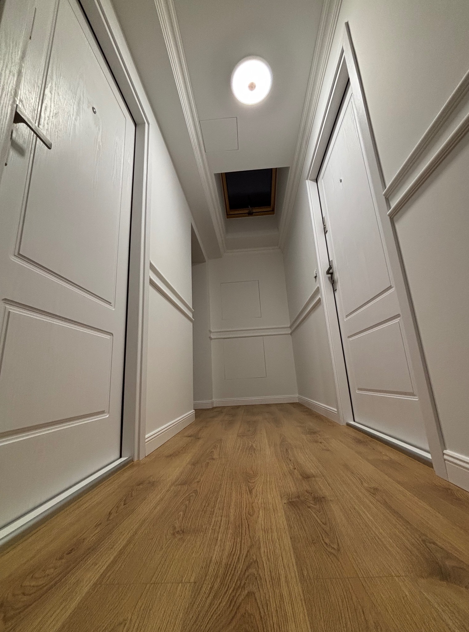 Biały korytarz z drewnianą podłogą, oświetleniem sufitowym i dwoma białymi drzwiami po bokach. Ujęcie z niskiej perspektywy podkreśla głębię i symetrię wnętrza.