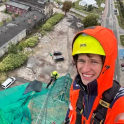 Uśmiechnięty pracownik w kasku i kombinezonie, na dachu zabezpieczonym siatką, z widokiem na miasto i drugiego pracownika przy pracy. Praca na wysokości.