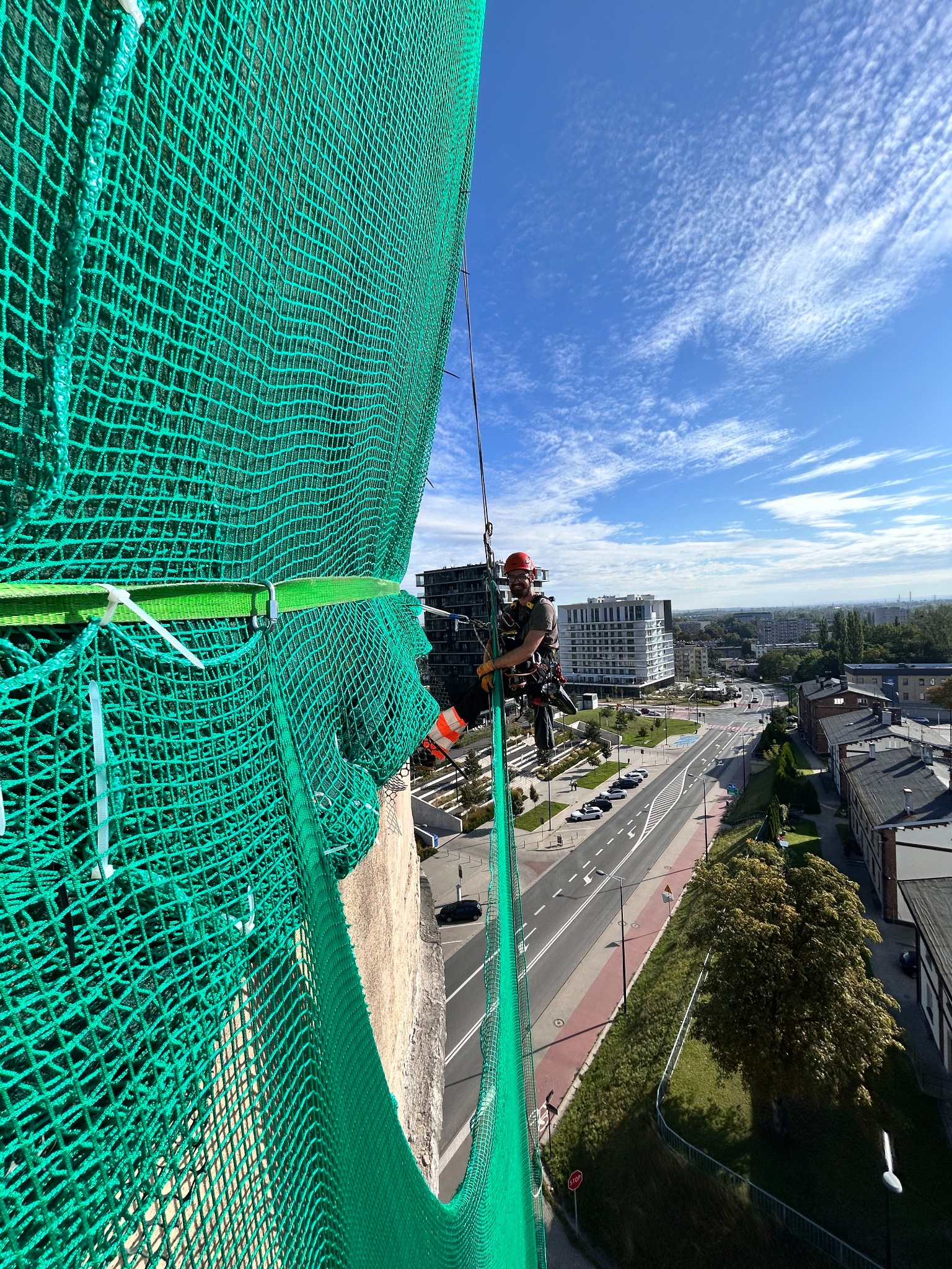 Pracownik w kasku, zabezpieczony linami, na wysokości budynku z zieloną siatką ochronną. Widok na miasto w tle, błękitne niebo z chmurami.