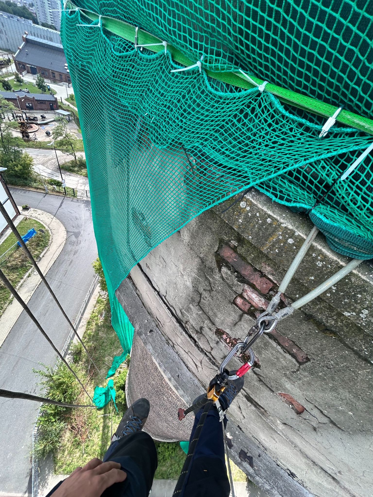 Praca na wysokości: widok z góry na zabezpieczoną siatką konstrukcję i osobę w uprzęży, z widocznymi elementami mocowania lin. W tle zabudowa miejska.