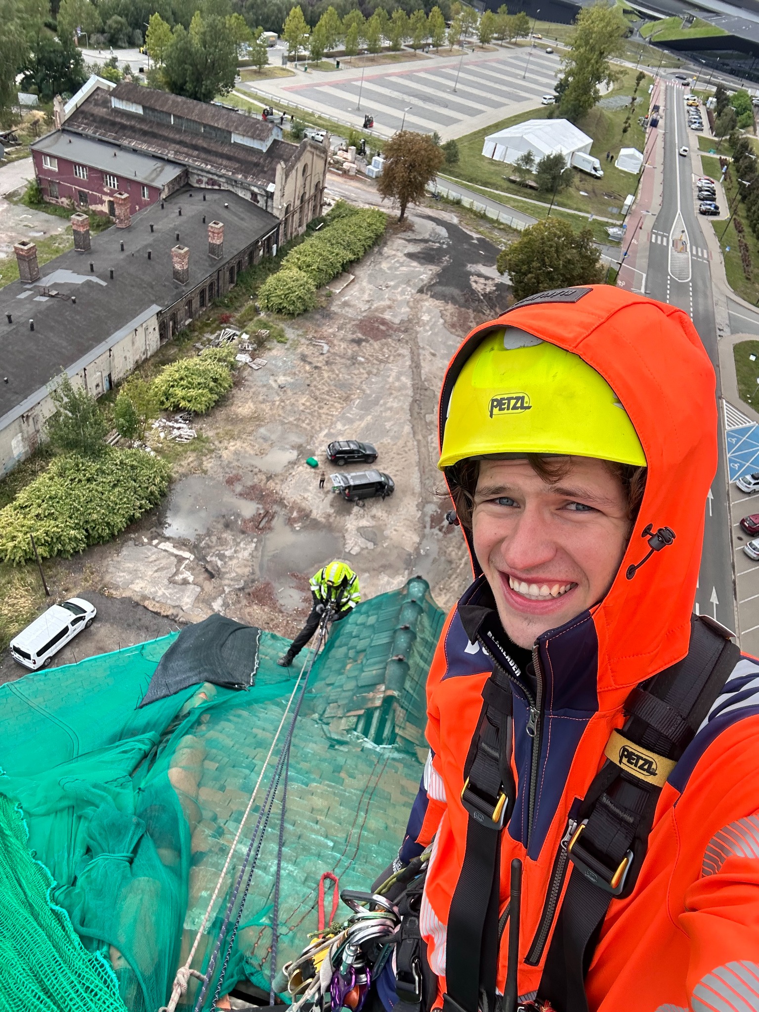 Uśmiechnięty pracownik w kasku i kombinezonie, na dachu zabezpieczonym siatką, z widokiem na miasto i drugiego pracownika przy pracy. Praca na wysokości.
