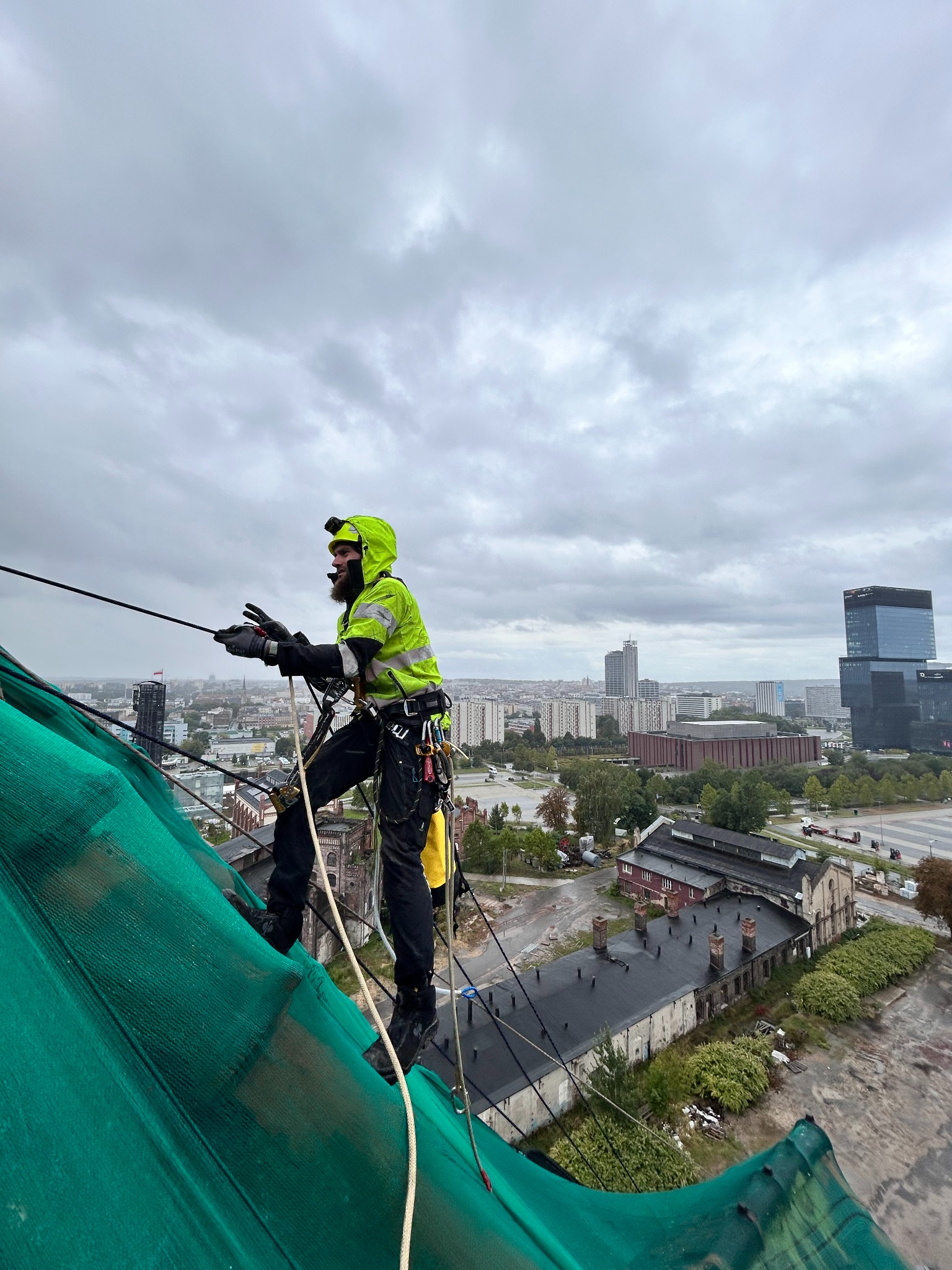 Pracownik w jaskrawej kurtce na wysokości, zabezpieczony linami, kontroluje stan dachu z zieloną siatką na tle panoramy miasta w pochmurny dzień. Prace wysokościowe.