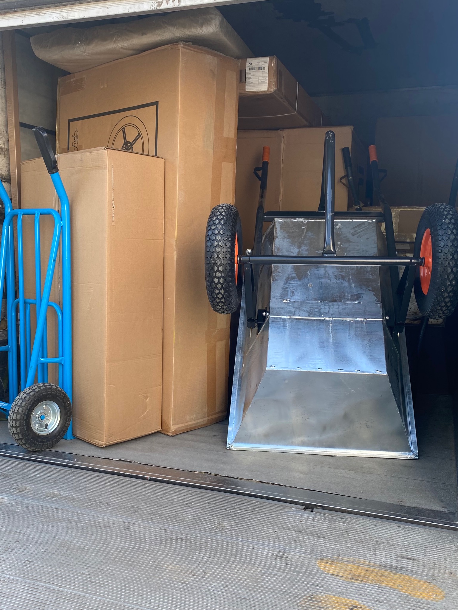 Wnętrze pojazdu transportowego z kartonami, niebieskim wózkiem transportowym i metalową taczką dwukołową. Widok na załadunek, przygotowany do transportu.