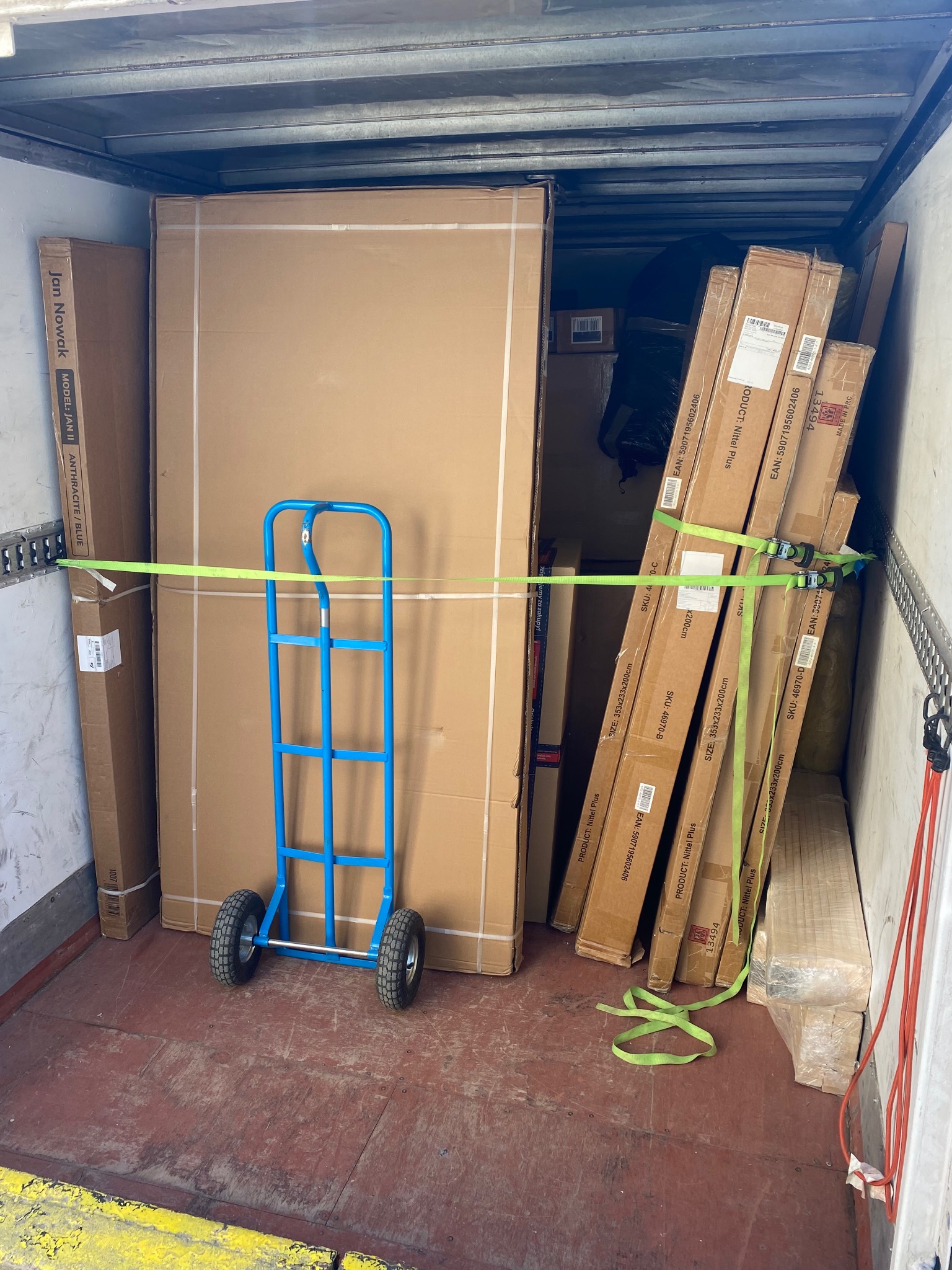 Wnętrze busa załadowane kartonami zabezpieczonymi pasami transportowymi. Na podłodze wózek transportowy. Towar gotowy do przewozu.