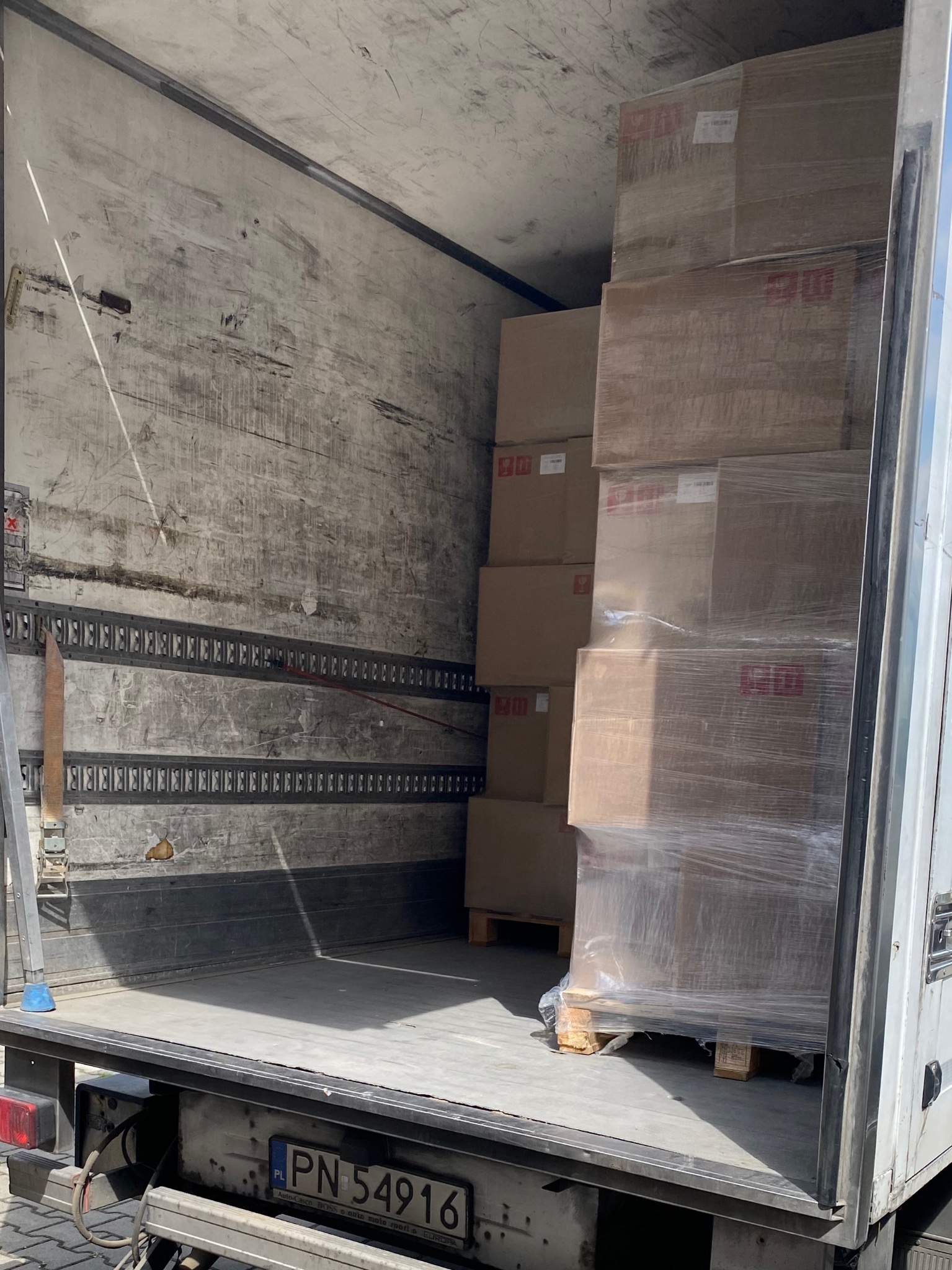 Wnętrze brudnej naczepy wypełnionej kartonami na paletach, zabezpieczonymi folią stretch. Widoczna tablica rejestracyjna pojazdu.