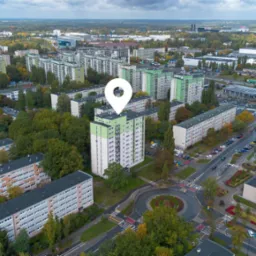 Panorama osiedla z lotu ptaka, zaznaczony budynek z zielonym dachem. Widoczne bloki, ulice, drzewa, parkingi i budynki użyteczności publicznej w jesiennej scenerii.