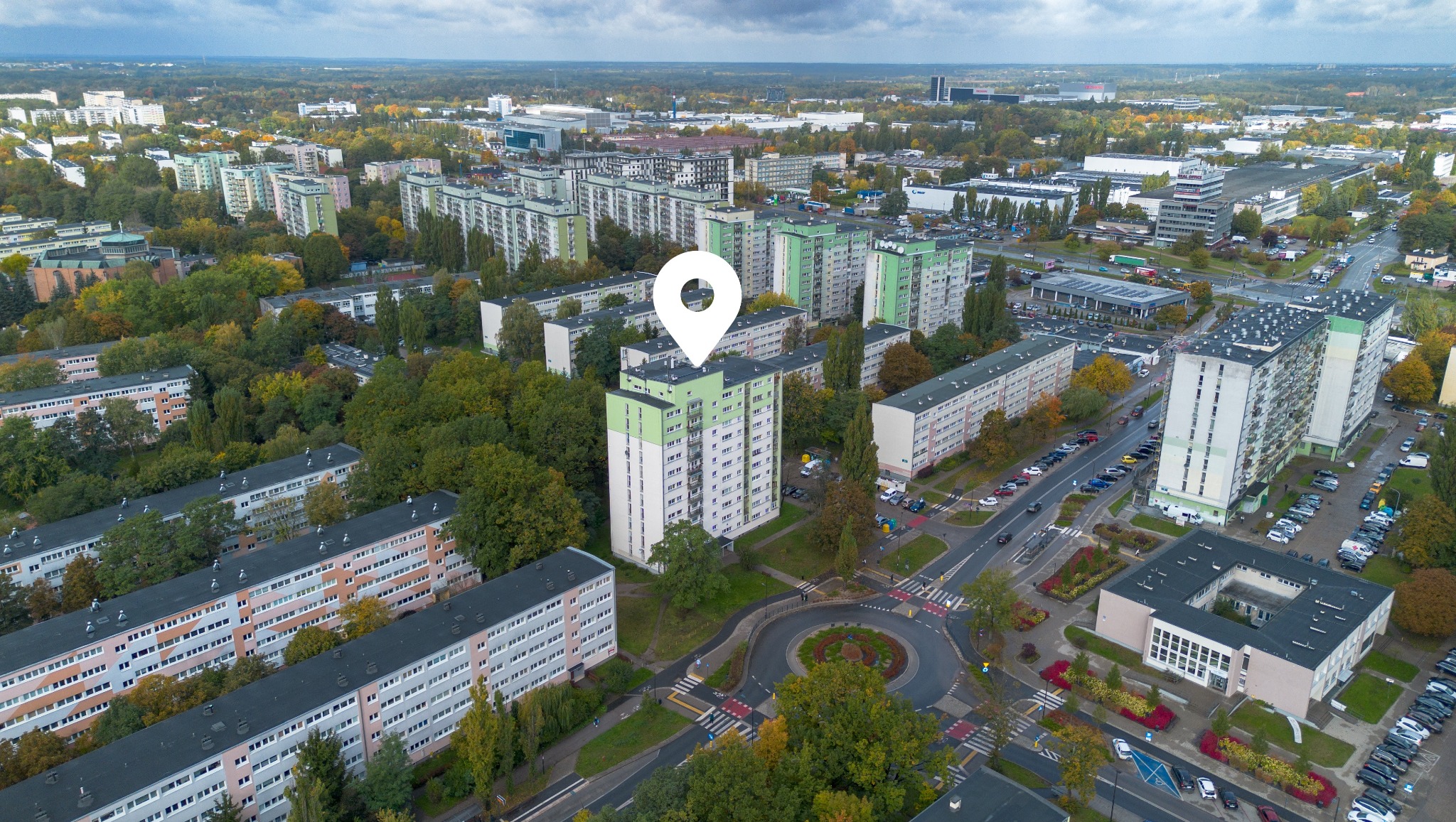 Panorama osiedla z lotu ptaka, zaznaczony budynek z zielonym dachem. Widoczne bloki, ulice, drzewa, parkingi i budynki użyteczności publicznej w jesiennej scenerii.
