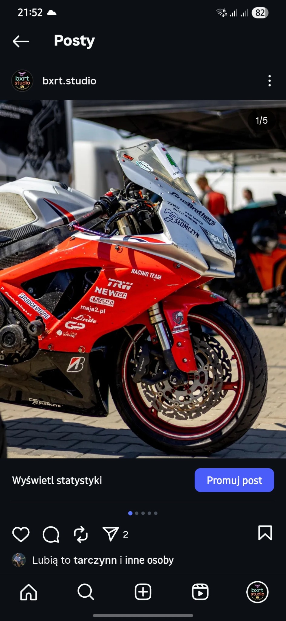 Czerwono-biały motocykl wyścigowy z widocznymi logotypami sponsorów, zaparkowany na zewnątrz, z detalami opon i układu hamulcowego.