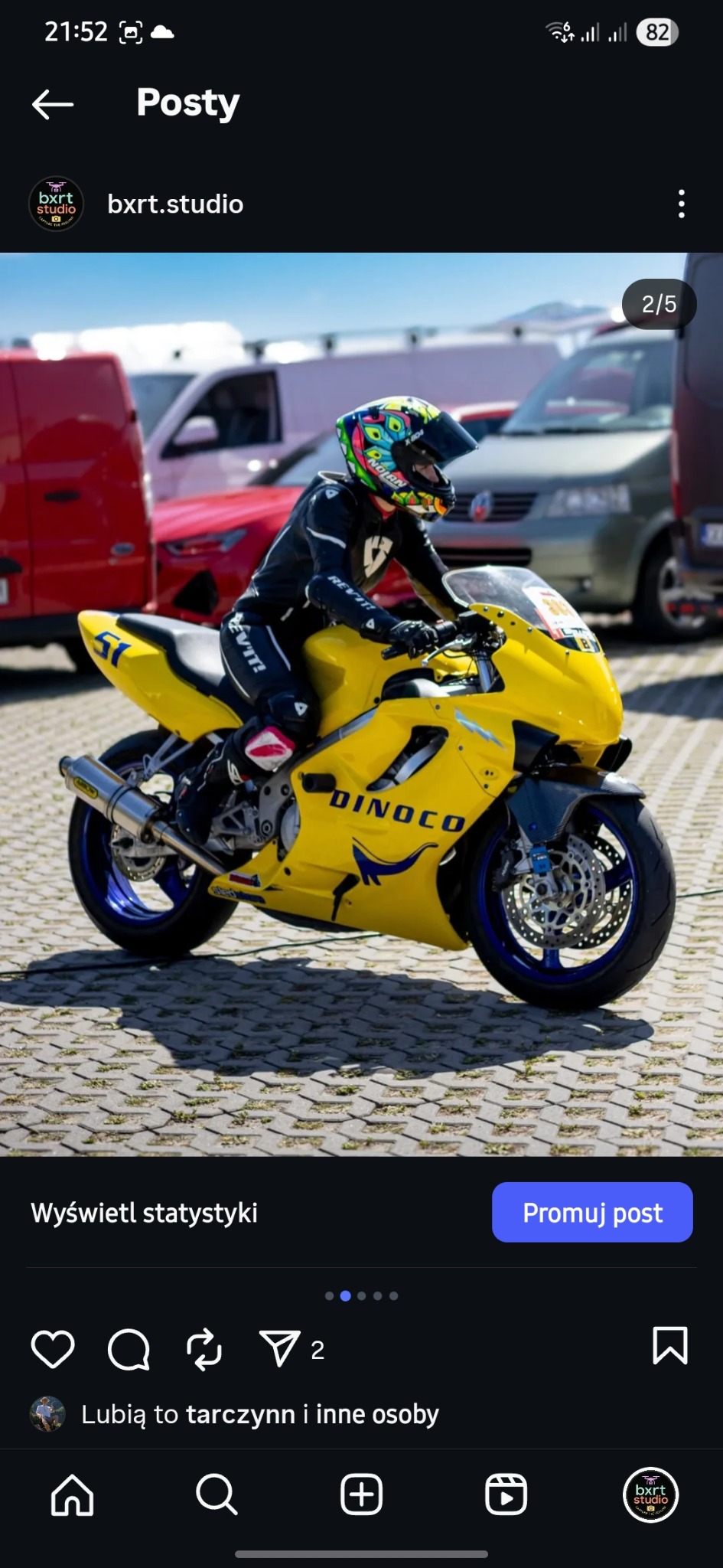 Żółty motocykl 'DINOCO' z niebieskimi felgami, na którym siedzi motocyklista w czarnym kombinezonie i kolorowym kasku. Tło stanowią zaparkowane samochody.